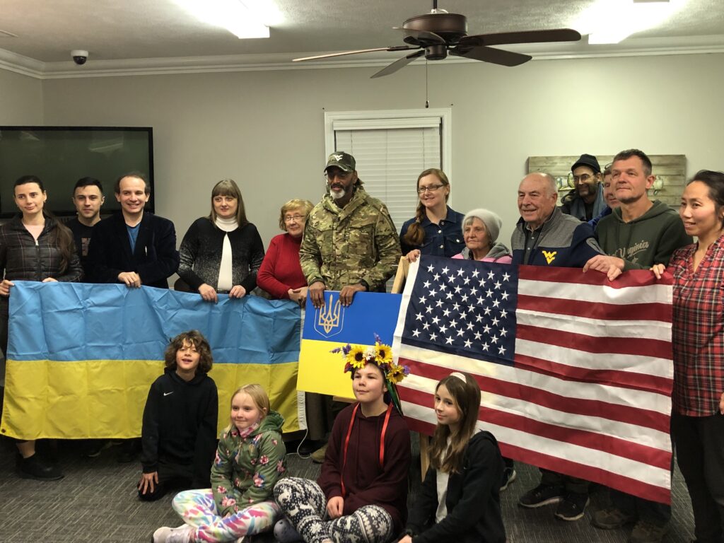 A group holds up a Ukranian flag on the left, as well as a United States flag on the right. A man in the middle of the group wears military fatigues. There is a group of children seated on the floor in front.