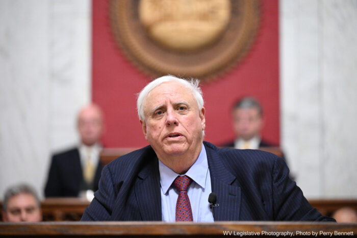 Gray haired man sits at a podium with two more men out of focus behind him.