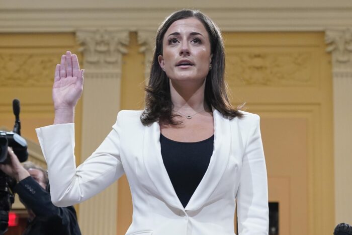 An adult woman raises her right hand during a public conference. She wears a white blazer and black shirt underneath.
