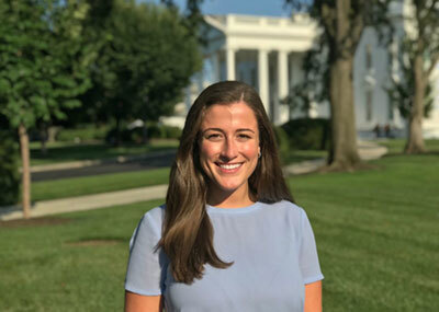 A young woman in a light blue shirt smiles for the camera. In the distance, is the White House.