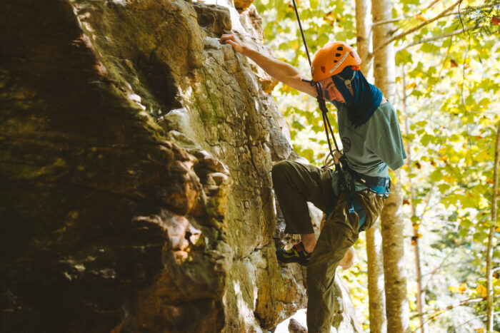 A young man climbs the side of a mountain. He wears a yellow helmet, blue t-shirt and khaki pants and climbing shoes. It's a sunny day.