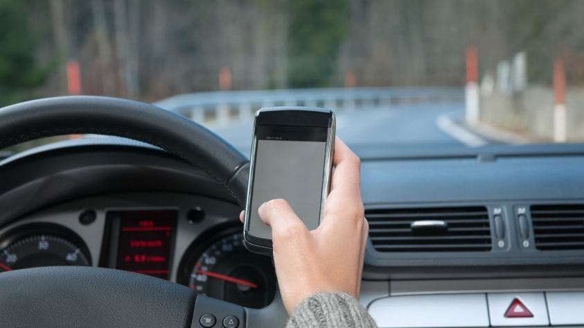 A hand can be seen holding up a cell phone in front of a car dashboard. Outside of the windscreen can be seen a road curving to the right.