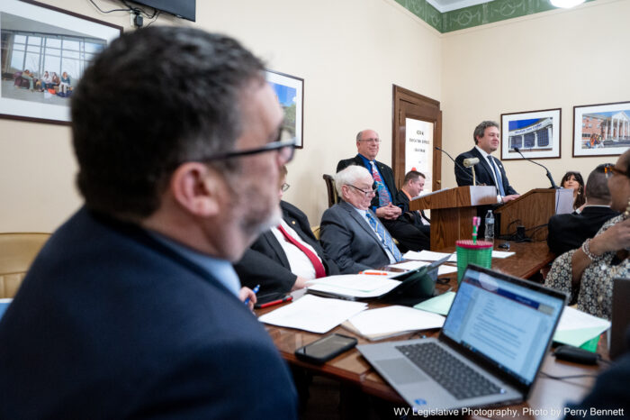 An out of focus man wearing glasses and a blue suit is in the foreground in a partial profile facing towards a speaker standing at a lectern. Between them are several people sitting at a wooden desk.