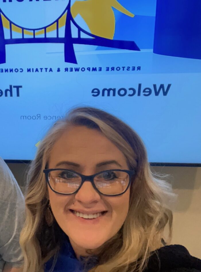 An adult woman with blonde hair takes a selfie. She smiles and wears glasses.
