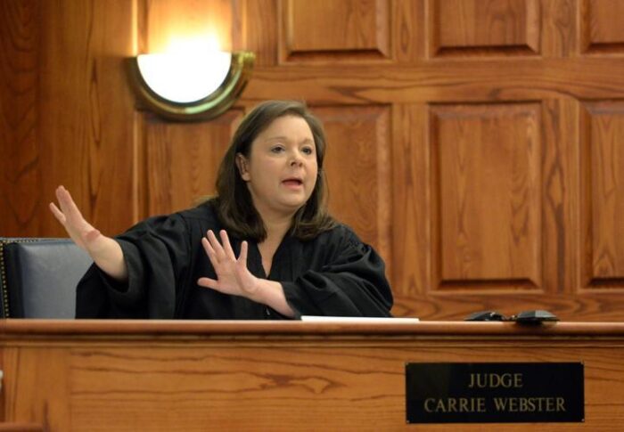 An adult woman, dressed in judge robes, sits at a judge's podium. She waves her hands to her right, as if in the middle of a phrase. A sign on the desk reads, "Judge Carrie Webster."