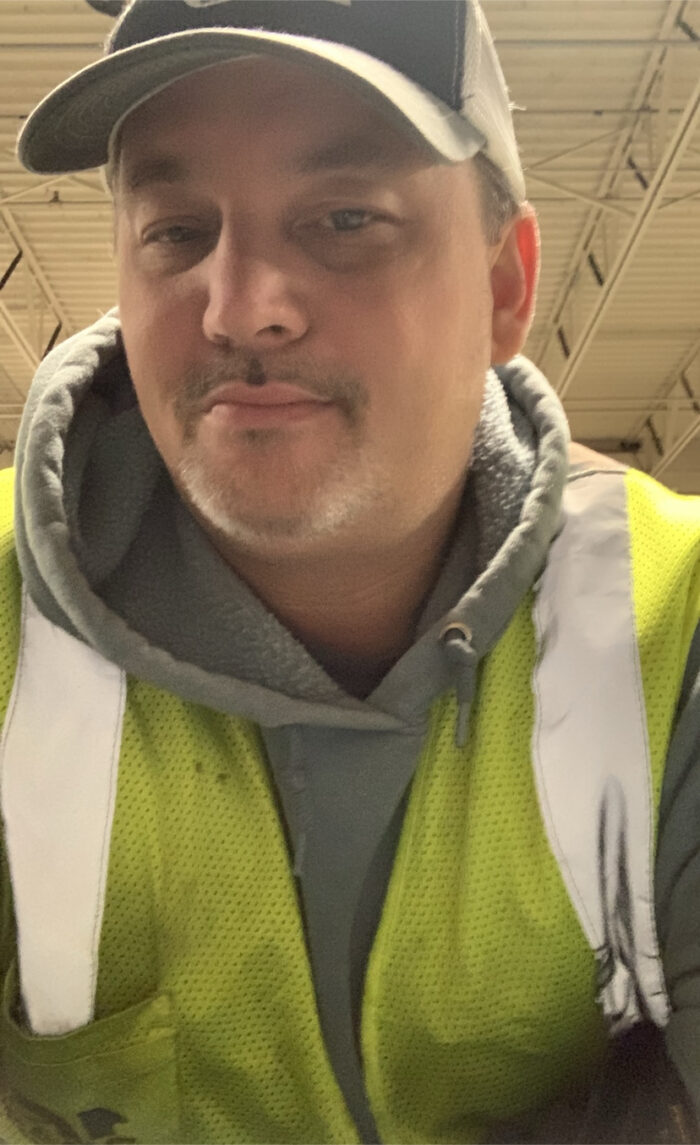 An adult man, wearing a ball cap and light reflector vest, takes a selfie.