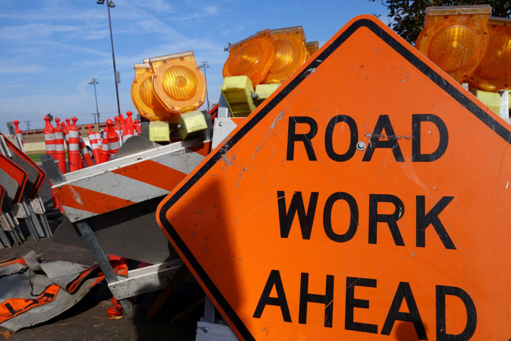 A neon orange sign that reads "Road Work Ahead" is positioned in front of barriers with reflectors.