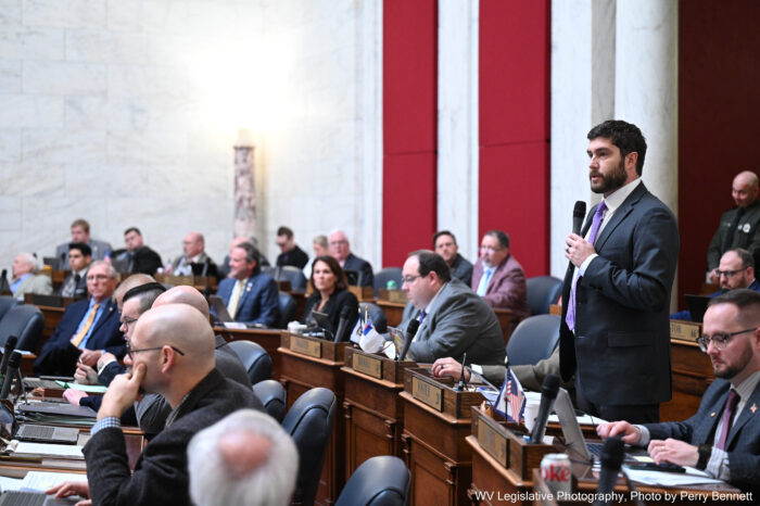 A man wearing a suit and tie speaks from his desk in a lawmaker's chambers. He uses a microphone.