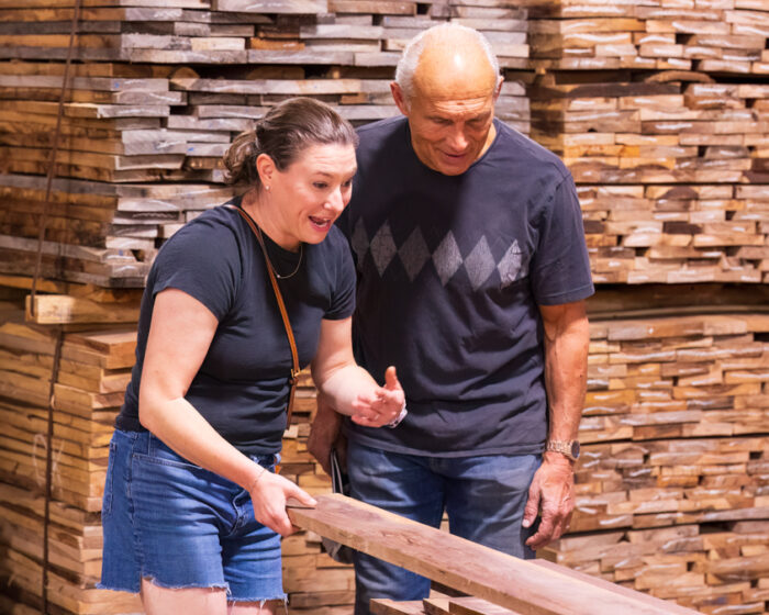 A middle age woman stands next to a balding man. There are stacked pieces of wood behind them.