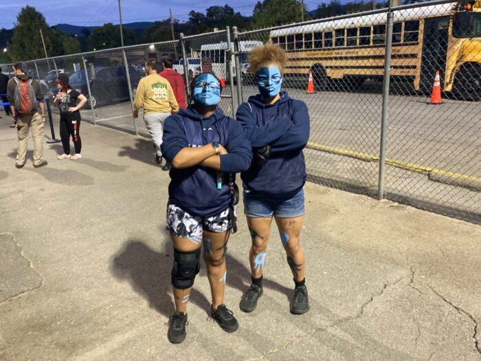 Two teen girls show off their face paint at a high school football game. One side of their faces is painted a dark blue, while the other side is painted a light blue.
