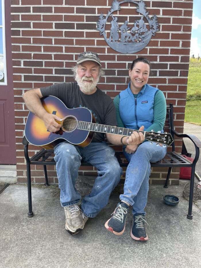 An older man sits on a bench next to a middle age woman. They both smile for the camera. The man is in a black t-shirt and jeans, and he holds a guitar. The woman is in a blue vest and long sleeved shirt, jeans, and sneakers.