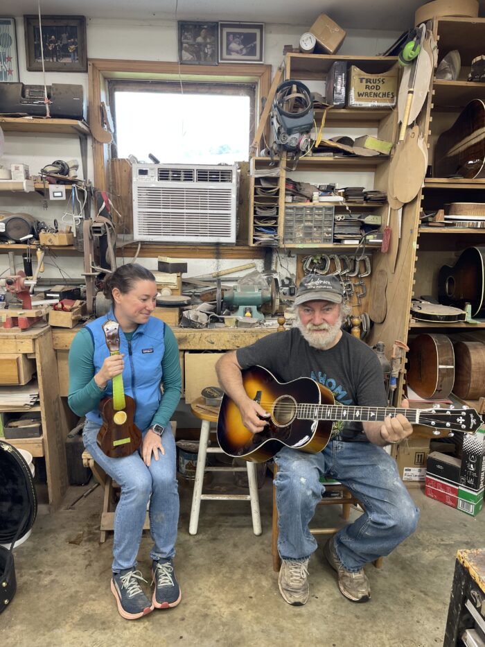 Two people - a middle age woman and an older man - sit next to each other smiling. The woman holds a ukulele and the man holds a guitar. They appear to be in a workshop.