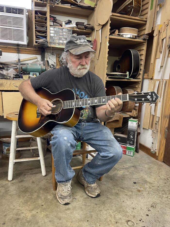 An older man sits in a chair dressed in jeans, a ball cap, and a t-shirt. He plays a guitar.
