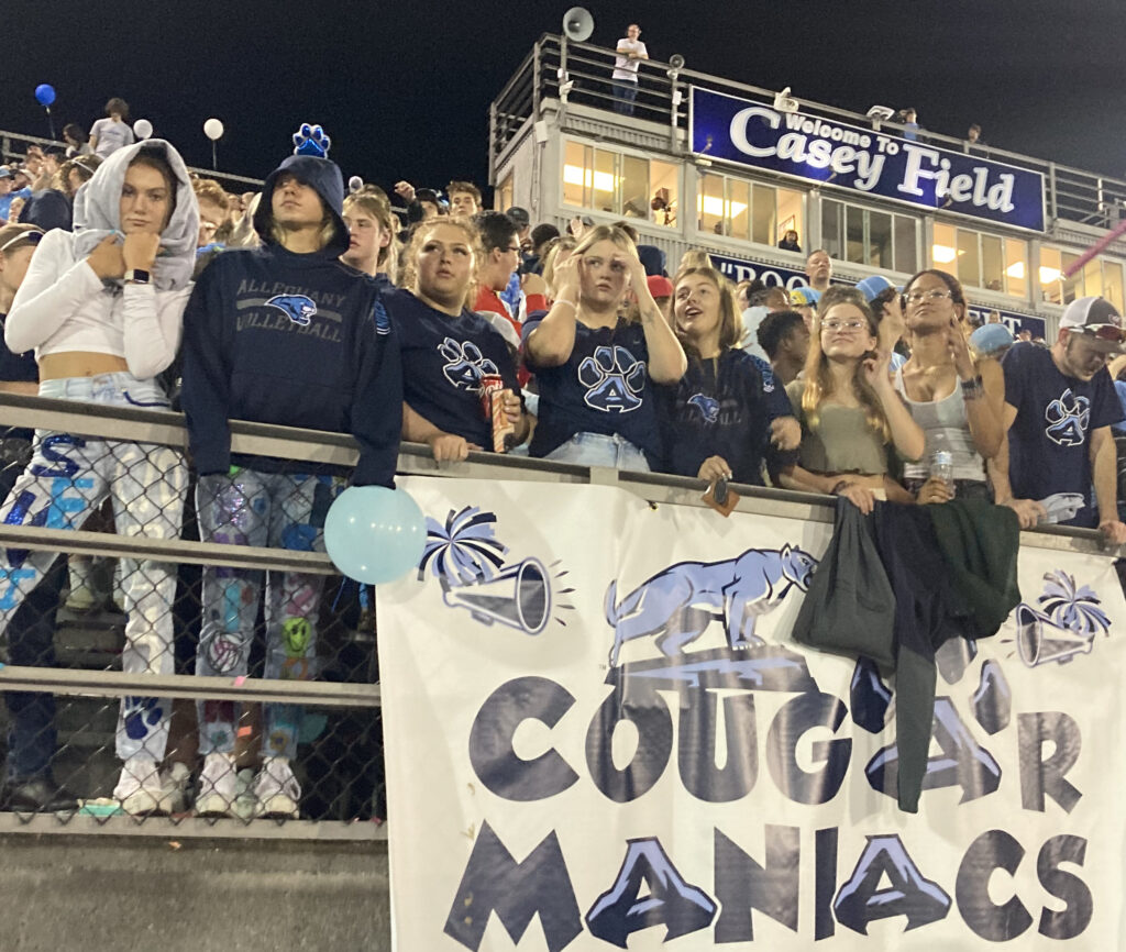 Dozens of teens attend a football game. In front of them, attached to the bleachers, is a sign that reads "Cougar Maniacs."