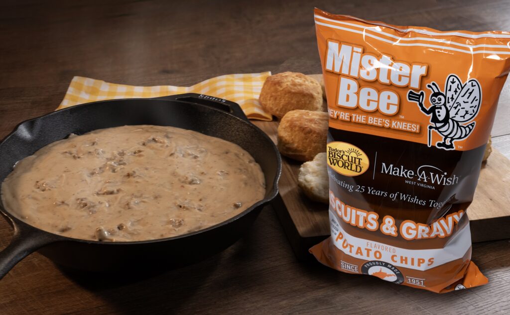 A skillet of sausage gravy sits next to a bag of Mister Bee Biscuits and Gravy flavored chips. Behind the chip bag can be seen several biscuits.