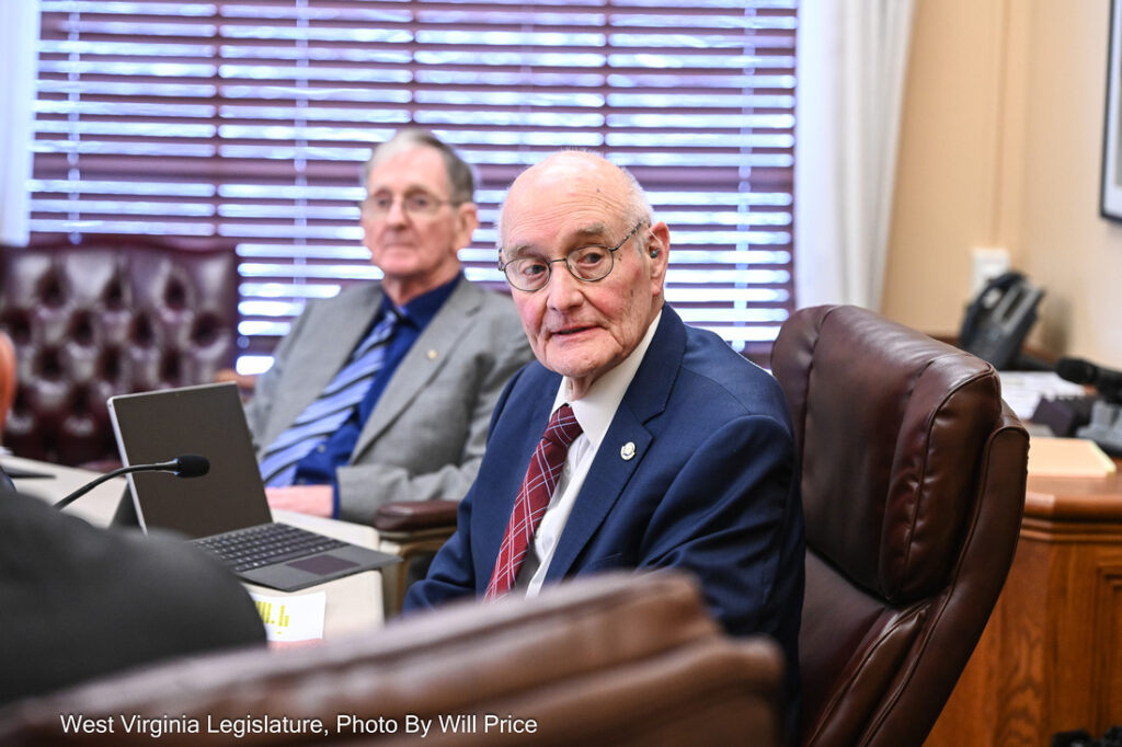 Senator Charles Clements sits at his desk at the Senate Transportation and Infrastructure Committee meeting, turned in his chair and glancing over his right shoulder. Another senator on the Committee sits behind him, slightly blurred.