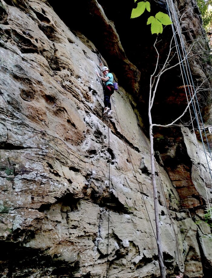 A woman climbs a rock wall. She smiles down at the camera.