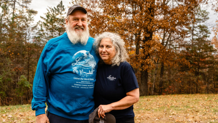 Two older people pose for a photo while giving each other a side hug. One individual is a man with a white beard, wearing a ball cap and blue long-sleeved shirt. The person next to him is a woman with gray, curly hair, wearing a black t-shirt. The leaves on the trees behind them are changing for fall.