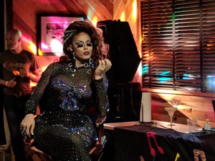 A drag queen inspects her nails, while sitting in a director style chair. She wears a beautiful black and blue sparkly dress.