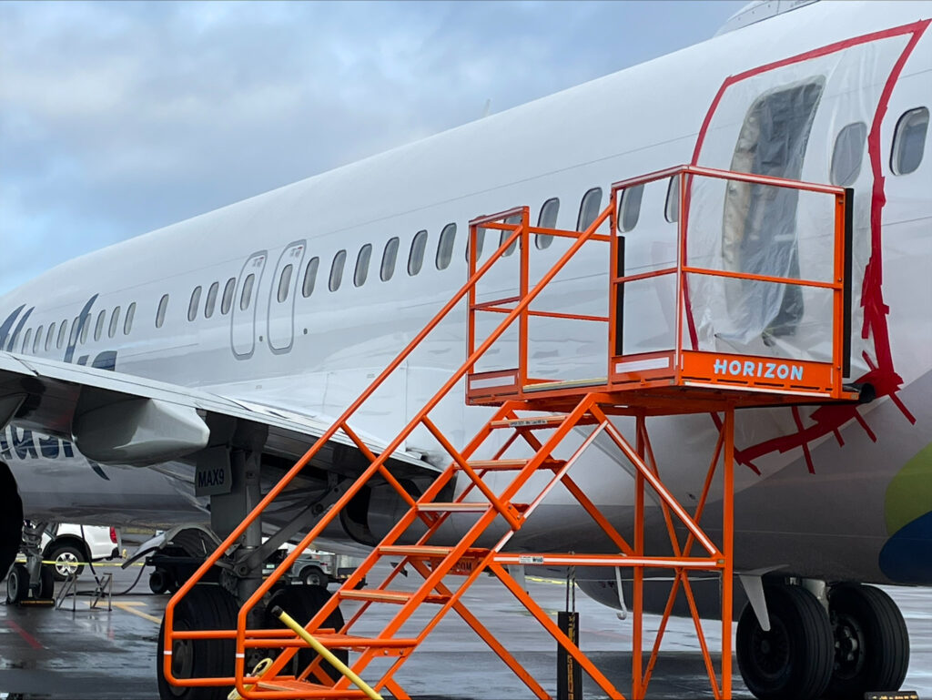 An airplane fuselage with a missing door is seen on a tarmac, with the door space covered in plastic.