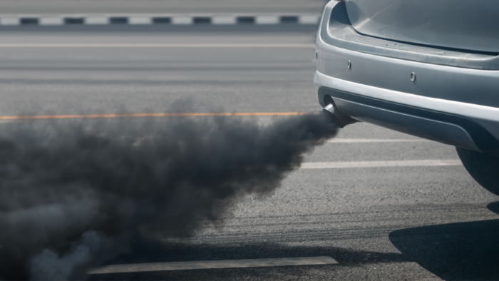 A black cloud of car exhaust is shown coming out of a car tailpipe along an asphalt road.
