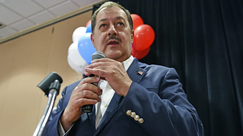 An older man speaks into a microphone, as red, white, and blue balloons are seen behind his head.