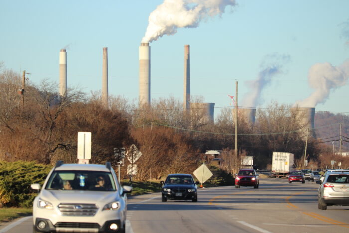 Wisps of power plant emissions rise into a clear sky as traffic passes on a highway in the foreground.