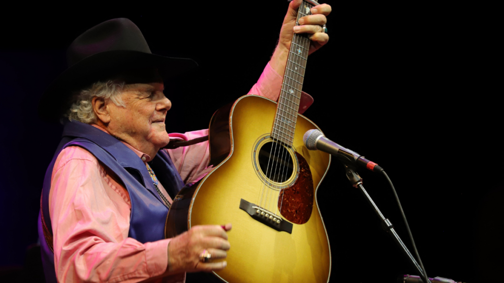 An elderly man with a black cowboy hat raises his guitar to the microphone.