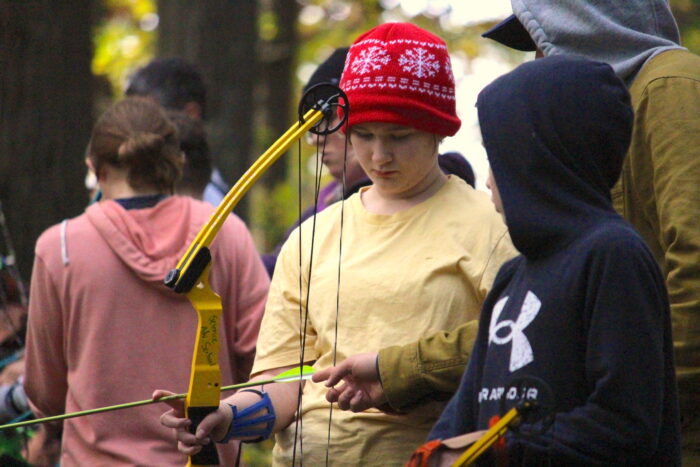 A student wearing yellow shirt and red cap stands next to another student wearing a dark blue UnderArmour sweater. Both are looking at compound bows in their hands. Behind them can be seen more students, as well as trees.