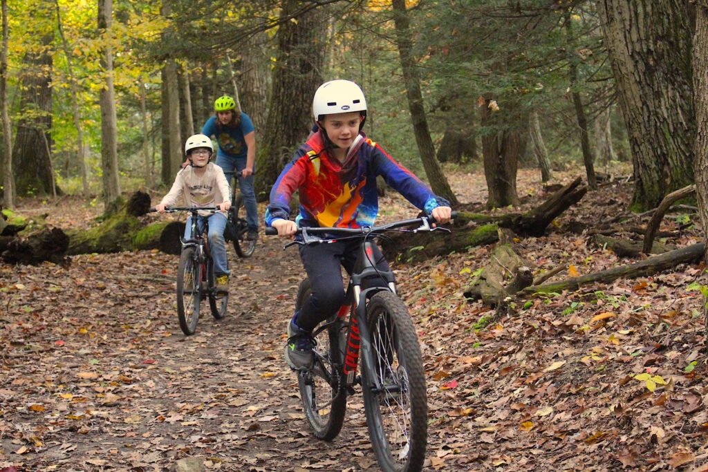 A student wearing a tie-dye sweater and a white helmet rides a bicycle through a forest. He is followed by another student, also wearing a white helmet and white sweater, as well as an adult in a short sleeved t-shirt and a lime-green helmet.