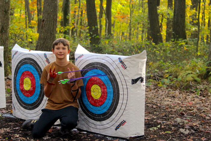 A student wearing a brown shirt kneels and puts up two peace signs next to an archery target with arrows sticking out of it. Three of the arrows are in a tight grouping in the target's blue ring, with a fourth arrow just above in the black. The archery setup is in a heavily forested area.
