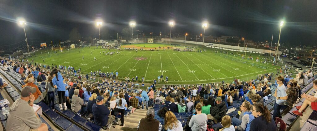 A wide photograph of a crowded football stadium lit brightly on a game night.