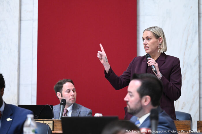 A woman stands speaking with a microphone holding one hand up.