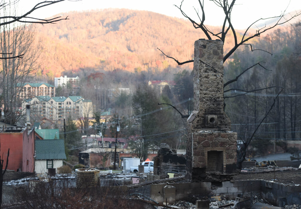 A charred, stone chimney is shown after a fire. In the distance, the sun appears to be setting, and there's a town surrounded by trees.