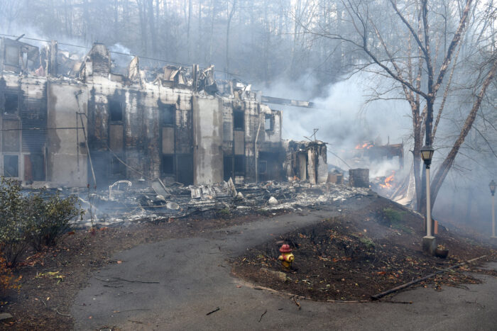 A building is almost indistinguishable after a devastating wildfire. The smoke billows, and there are flames in the distance. A single lamppost appears untouched.