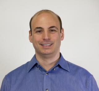 A photo of a balding man wearing a blue button up shirt. He's smiling and posing for a photo.