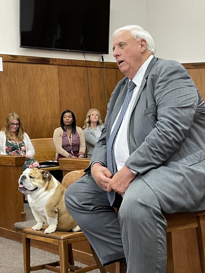 A large man sits in a chair wearing formal attire. Next to him, in a lower chair, is an English Bulldog.