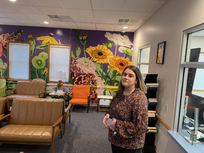 A young woman stands in a colorful waiting room. There are flowers painted on the wall.
