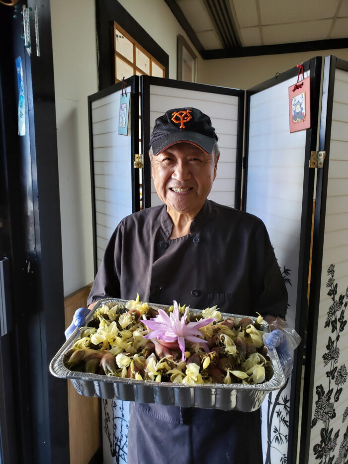 An older Japanese man smiles for the camera while holding a tray full of food. He wears a black ball cap.