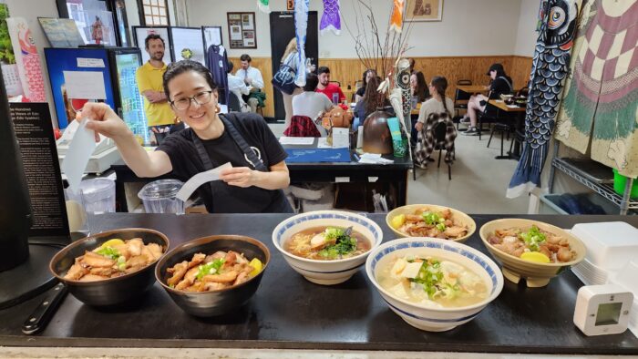 A woman takes an order. In front of her is a line of delicious looking Japanese food on a counter. She smiles for the camera and wears a black apron.