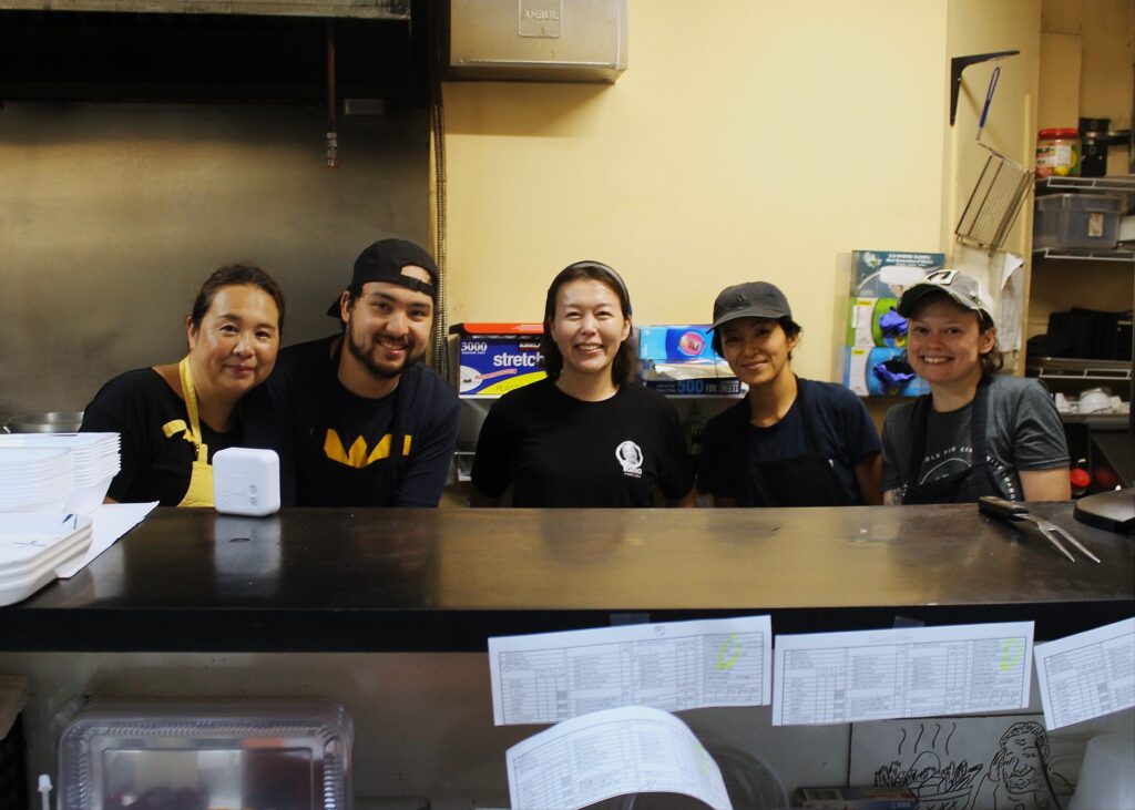 Six smiling people are shown - five women and one man. They pose for a photo from behind a counter at a restaurant they work at. Some are dressed with black aprons.