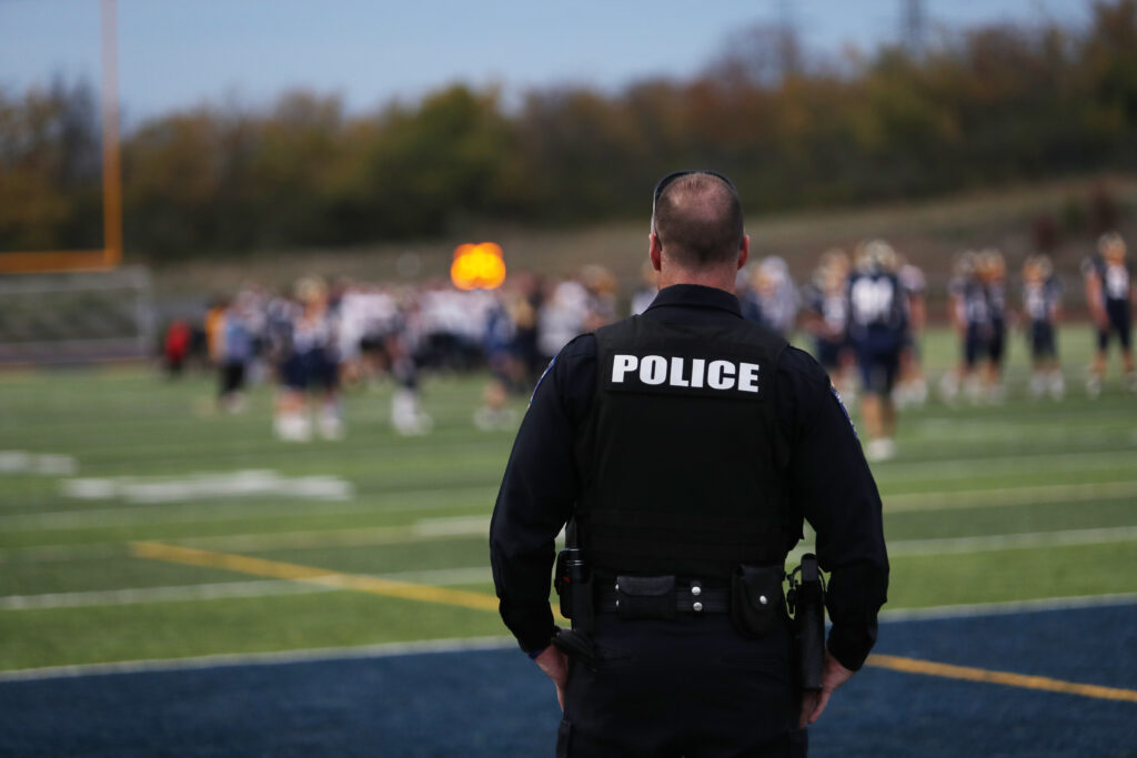 A police officer dressed in black and the word POLICE across his shoulders in white stands in the endzone of a football field facing away from the frame. In the background, out of focus, can be seen football players in blue and white.