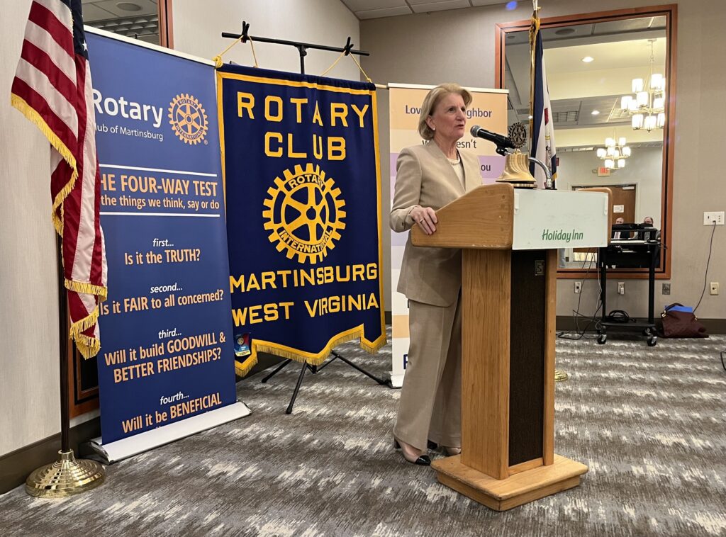 Senator Shelley Moore Capito stands at a podium and speaks into a microphone. Behind her are the American flag, and a poster for the Rotary Club of Martinsburg.