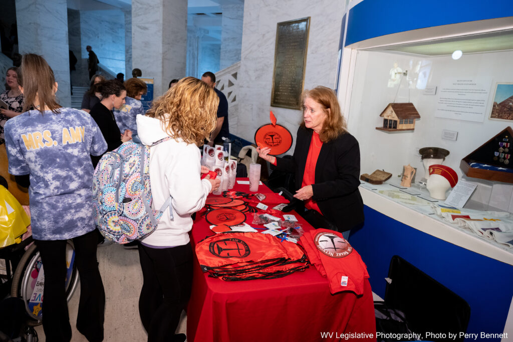 A woman wearing a black blazer over a red shirt stands at a table covered in a red tablecloth. The table is covered in materials emblazoned with the logo of Fair Shake West Virginia. On the other side of the table stands a person wearing a white sweater and a backpack.
