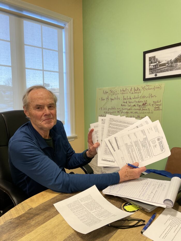 An older man sits at a desk holding several papers up to the camera. He has gray hair and is wearing a dark blue long-sleeved shirt.