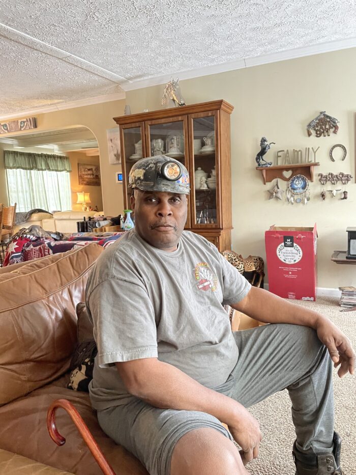 An older man wearing a miner hat sits on the couch of his home and poses for a photo. Next to him, leaning against the couch is a brown, wooden cane.