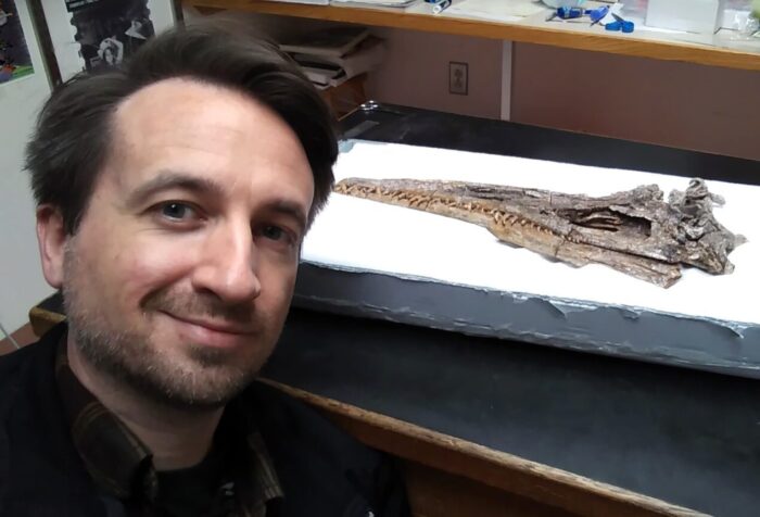 A man with dark hair poses with a fossilized skull of a plesiosaur.