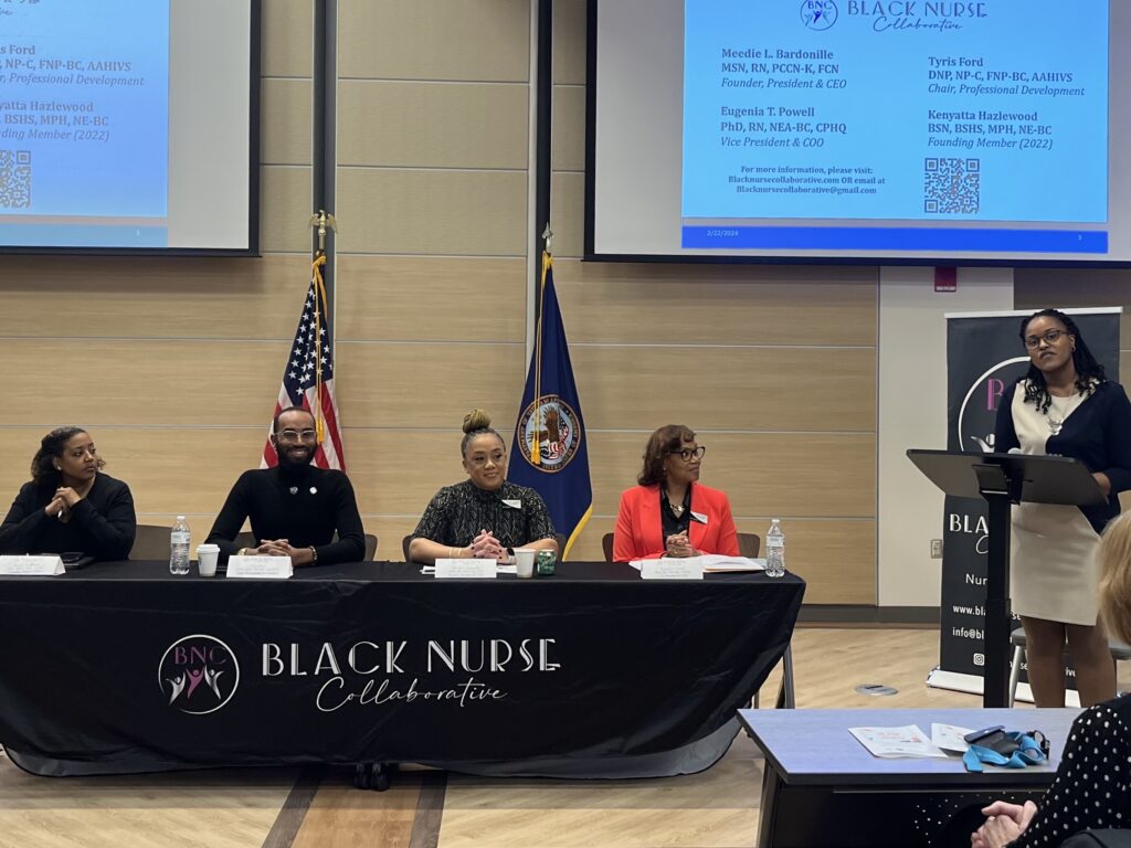 Four panelists sit at a table with a tablecloth that says "Black Nurse Collaborative." They are looking to their left, toward a speaker at a podium who is introducing them to an audience off screen.