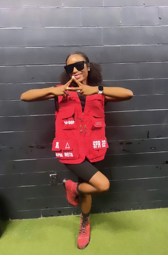A young woman poses for a photo. Her hair is dark and pulled back. She wears sunglasses, a red vest, black shorts, and red shoes.
