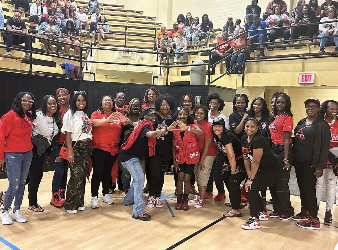 More than a dozen people pose for a photo on a basketball court. They're smiling, wearing mostly red, black, or white shirts. All are women.